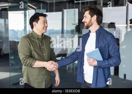 Two smiling multiethnic businessmen shaking hands, making a professional greeting and showing their partnership and collaboration in a modern office meeting Stock Photo