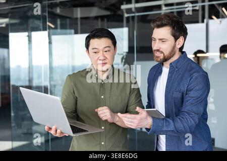 Diverse colleagues collaborating over laptop and tablet, discussing data and strategy in a modern office, sharing ideas for project planning, training, and business growth Stock Photo