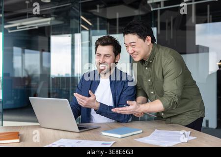 Diverse business colleagues smiling and looking at a laptop screen in a modern office, engaged in teamwork and discussing a project, showing successful collaboration Stock Photo