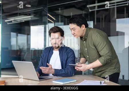 Two diverse business colleagues working together, discussing a project while looking at a laptop in a modern office, showing teamwork and professional collaboration Stock Photo