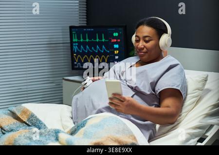 Pregnant woman resting in hospital bed, using smartphone while monitoring baby heartbeat and contractions. Ideal for maternity care, telehealth, prenatal support marketing Stock Photo