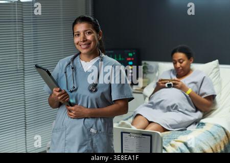 Portrait of Latin nurse holding tablet and standing beside pregnant woman using smartphone in hospital room. Supporting prenatal care Stock Photo