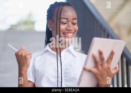 Happy young Black woman celebrating while holding tablet and stylus on stairs. Achievement, success, online learning, business progress, digital produ Stock Photo