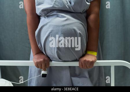 Unknown pregnant woman gripping hospital bed rail while standing in maternity ward, showing labor pain and childbirth care. Useful for prenatal healthcare, delivery services Stock Photo