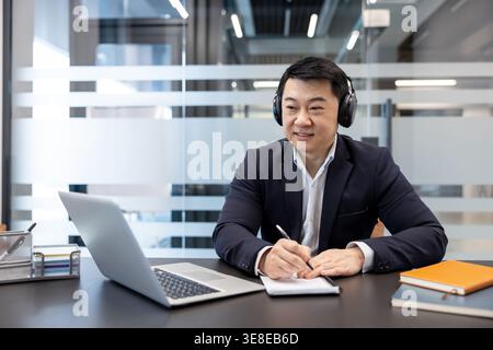 Smiling asian businessman wearing headphones, attentively listening and taking notes during a virtual conference or remote work session in a modern office environment Stock Photo