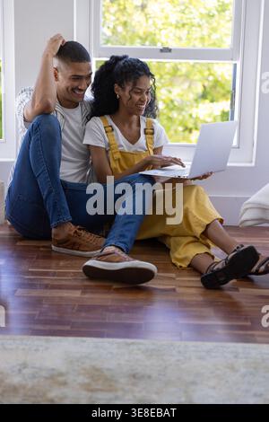 African American couple sitting at home, woman yellow overalls typing on laptop man leaning smiling Stock Photo