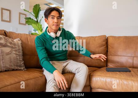Asian man sitting on tan leather sectional couch wearing white headphones, reaching for tablet Stock Photo