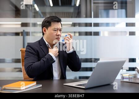 Asian businessman having a sudden asthma attack in the modern office, holding an inhaler and pressing his hand to his throat while working on a laptop Stock Photo