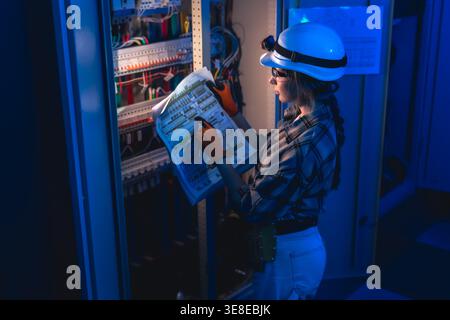Electrician inspecting electrical switchboard using technical blueprint. Industrial energy system. Stock Photo