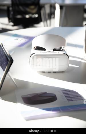 White VR headset resting centered on meeting table, left tablet and printed charts showing graphs. Device, daylight, shadows, glossy, modern, boardroom, office Stock Photo