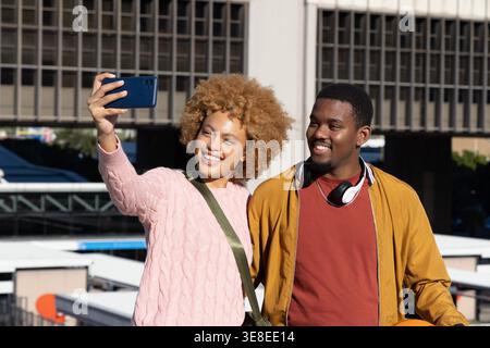 African American couple taking selfie with smartphone on rooftop near office tower with basketball. Urban, outdoor, sunny, candid, duo, smiling, terrace Stock Photo