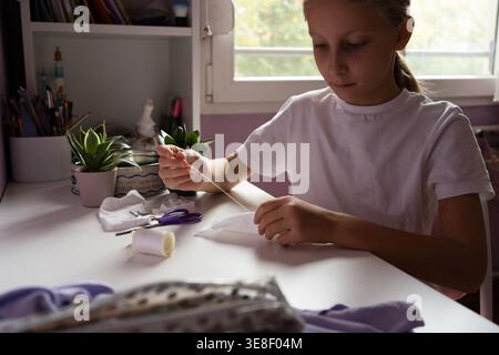 A girl sits at a white table sewing white fabric with a needle and thread surrounded by scissors and spools in a bright room with plants and bookshelf Stock Photo
