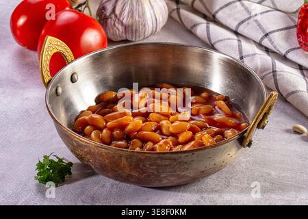 A close up of baked beans in a rustic metal bowl. Surrounded by fresh ingredients like tomatoes, garlic, and parsley on a grey table. This warm, appet Stock Photo