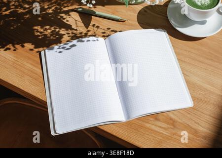 Close-up overhead view of an open notebook with blank pages and a hot matcha latte drink on a wooden table with floral shadows Stock Photo