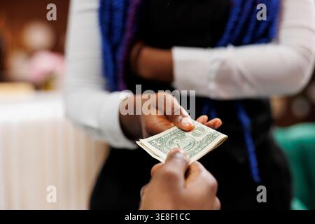 Close up of hotel employee wearing uniform holding money cash taking payment from customer while working in hospitality industry, selective focus. Waitress receiving tip from generous client Stock Photo