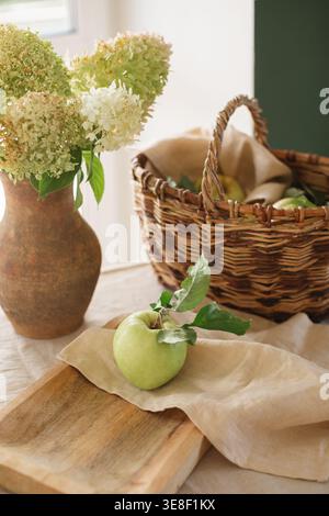 Bunch of fresh hydrangeas in a rustic clay jug on a table with a basket and an apple on a wooden chopping board with a napkin Stock Photo