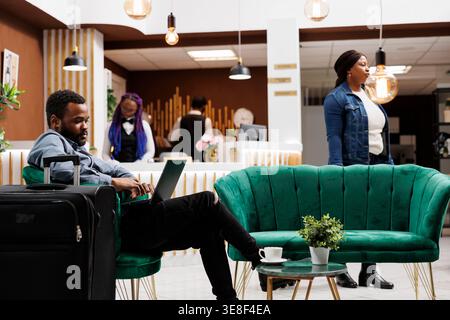 Busy African American man travelling freelancer sitting in armchair with suitcase and laptop at hotel lobby. Black millenial guy using computer working on project remotely during business trip Stock Photo