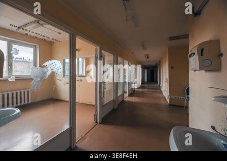 Interior view of a long hallway in a derelict Soviet children's hospital. Rows of empty glass-walled wards with faded teddy bear decals. Dusty floors Stock Photo