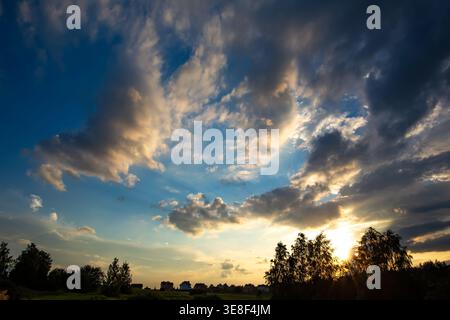 A vibrant wide-angle shot captures a partly cloudy sky transitioning from deep blue to bright hues, with the sun illuminating clouds in warm yellow Stock Photo