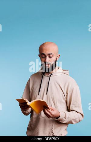 Arab man reading book paperback edition with yellow cover. Young concentrated bald bearded person in hoodie holding open notepad while standing with surprised facial expression Stock Photo
