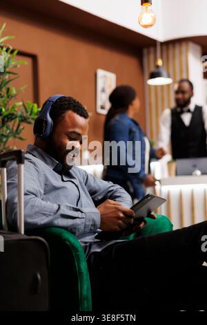 Smiling African American guy traveler sitting with luggage in hotel lobby, male tourist holding digital tablet connecting internet wifi while waiting for room to be ready. Guest self check-in Stock Photo
