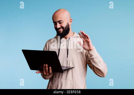 Arab man holding laptop while talking on video call, smiling and waving hello. Cheerful person having remote communication using portable computer app and greeting customer Stock Photo