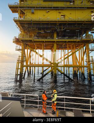 Two offshore workers in PPE converse on vessel deck alongside a large yellow offshore platform, North Sea, United Kingdom Stock Photo