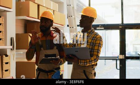 Employees team checking goods and cargo in storehouse, using laptop and clipboard list in depot. Two men organizing products in cardboard packages on racks, check merchandise. Stock Photo