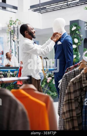 Clothing store assistant examining suit on mannequing and fixing tie to display new collection. Fashion boutique african american man employee dressing model in stylish formal menswear Stock Photo