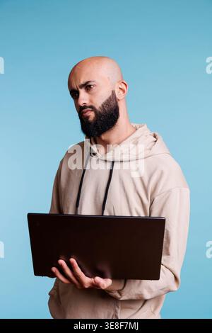 Puzzled arab man programming on laptop and looking away with confused expression. Confused person in doubt while developing software app and writing code on portable computer Stock Photo