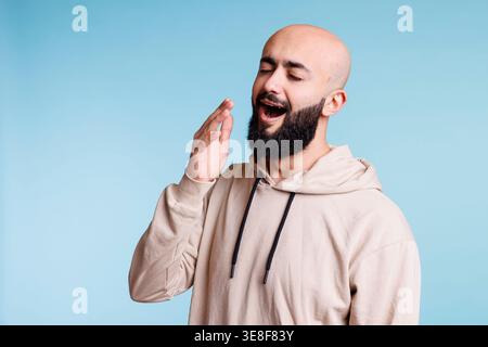 Sleepy young adult arab man with closed eyes yawning and covering mouth with hand. Tired bald bearded arabian person wearing casual beige hooding feeling exhausted and bored Stock Photo
