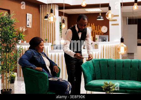 African guy hotel waiter serving hot aromatic coffee to woman guest sitting at lobby. Black woman traveler ordering cappuccino while waiting for check-in at reception area, Caffeine and jet lag Stock Photo