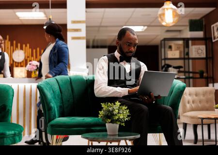 Focused African American man hotel manager sitting in lobby working on laptop, looking at computer screen, keeping track and managing bookings. Software for hospitality industry Stock Photo