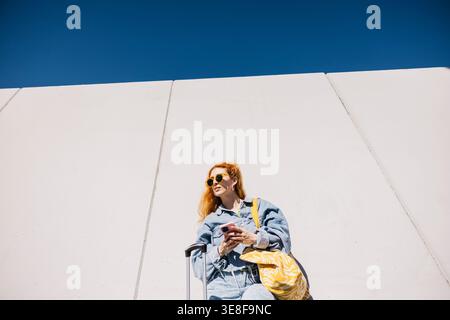 Woman standing with a suitcase and bag, using smartphone against a white wall and blue sky Stock Photo