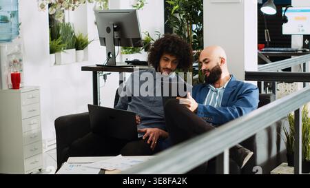 Two employees comparing notes in office, reviewing online company data on laptop and tablet. People planning new presentation for executive board meeting, using official papers. Tripod shot. Stock Photo