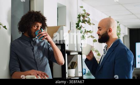 Coworkers joking around on coffee break, having conversation and laughing. Company employees relaxing in open floor office talking together, modern partnership in coworking space. Stock Photo