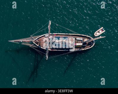 Aerial view of a large wooden sailing vessel with solar panels on deck and a small inflatable dinghy alongside on deep blue water. Stock Photo