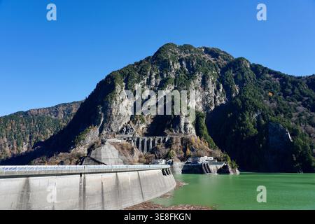 Concrete Dam and Reservoir Nestled Below Rugged Mountain Landscape Under Clear Blue Sky Stock Photo