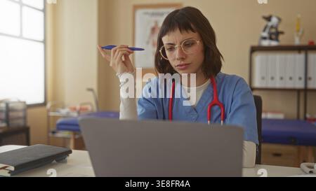Woman holding pen at laptop in clinic with red stethoscope hanging on neck and anatomical chart behind; concentration. Stock Photo
