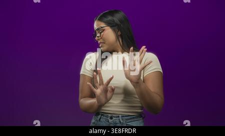Young woman with glasses and hands raised, recoiling with a disgusted facial grimace, wearing beige tee and jeans in a purple studio; disgust avoidanc Stock Photo