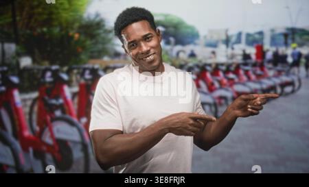 Man points with both hands toward a row of red rental bicycles at a street bike share station while smiling; urban mobility confidence. Stock Photo