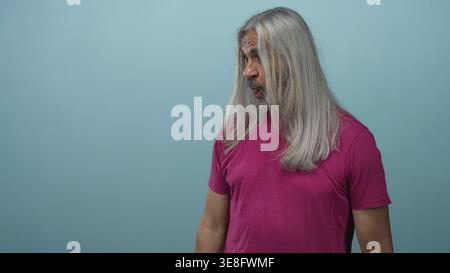 Middle aged man with long gray hair wearing magenta tee in teal studio looking sideways and talking with expressive face; surprise reflection. Stock Photo