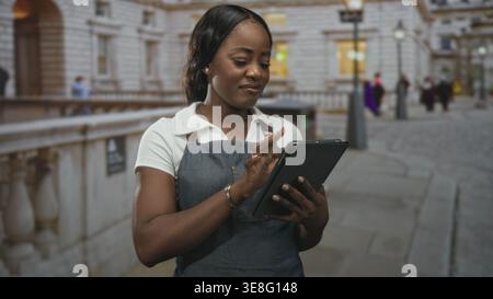 Woman in apron taps a tablet while holding it, smiling and checking orders on a cobblestone street outside a stone building; quiet focus service. Stock Photo