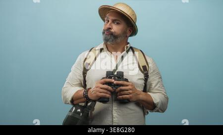 Middle aged hispanic man with grey beard holding binoculars, wearing backpack and pith helmet in blue studio; curiosity travel exploration. Stock Photo