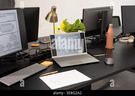 Silver laptop displaying floor plan on black desk in office with monitors, coral water bottle Stock Photo