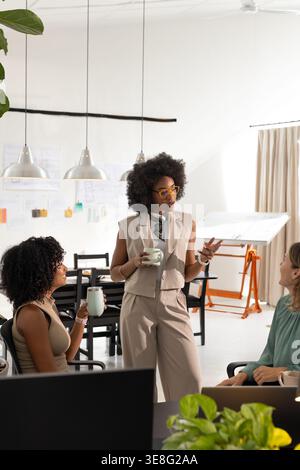 Diverse women team ideating sketches in studio, standing African American gesturing with mug Stock Photo