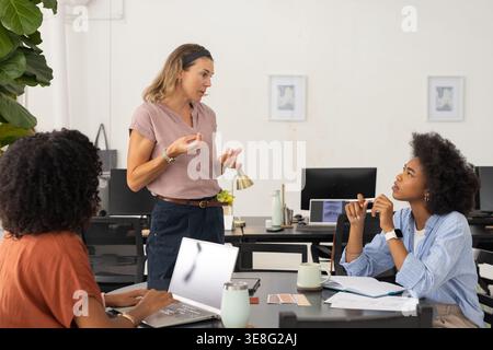 Diverse female coworkers discussing at work table in open office, using laptop and color swatches Stock Photo