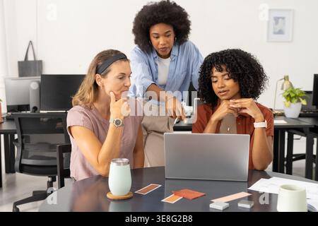 Diverse female coworkers collaborating around round table in bright office with laptop, swatches Stock Photo