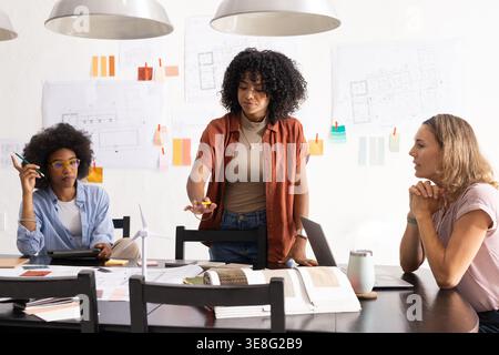 Diverse female coworkers studying blueprints at design studio, standing woman holding yellow object Stock Photo