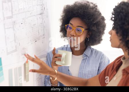 Diverse female designers studying pinned floor plans with color cards in studio, holding mug Stock Photo